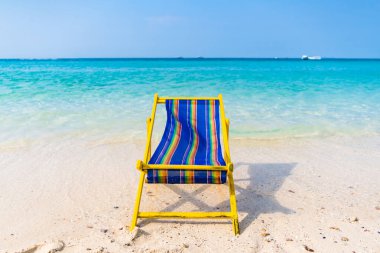 Berrak, turkuaz sulara ve berrak mavi gökyüzünün altındaki yemyeşil tepelere tepeden bakan el değmemiş beyaz kumsalda canlı, sarı ve mavi bir sahil sandalyesi bulunan sakin bir tropikal plaj sahnesi..