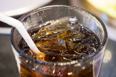 Buzlu bir bardak kola ve restoran masasında pipet ile yakın plan, çeşitli yemekler ve çatallarla çevrili, ortak bir yemek deneyimini ve içeceğin ferahlatıcı doğasını tasvir eden.