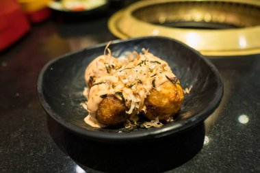 Çeşitli malzemelerle süslenmiş taze yapılmış Takoyaki topları. Lokanta masasındaki siyah bir kasede sıcak servis ediliyor..