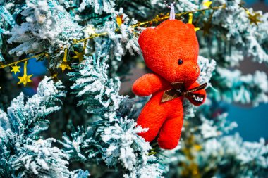 Bright red teddy bear ornament hanging on a snow-covered Christmas tree branch, surrounded by golden star garlands and festive holiday decor. clipart