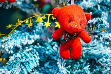 Karla kaplı bir Noel ağacının dalında asılı parlak kırmızı oyuncak ayı süsü. Altın yıldız çelenkleri ve bayram dekorlarıyla çevrili..