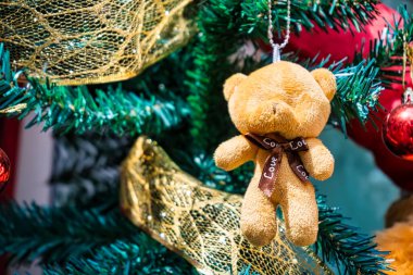 Altın kurdele ve canlı kırmızı mücevherlerle süslenmiş bir Noel ağacında asılı duran yumuşak kahverengi oyuncak ayı süsü. Bayram dekoruna sıcaklık ve cazibe katıyor..