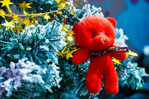 Karla kaplı bir Noel ağacının dalında asılı parlak kırmızı oyuncak ayı süsü. Altın yıldız çelenkleri ve bayram dekorlarıyla çevrili..