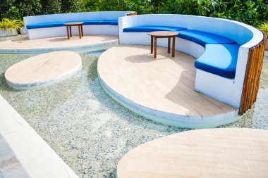 A contemporary outdoor seating area with curved wooden platforms, blue cushioned benches, and small wooden tables, surrounded by lush greenery for a relaxing ambiance. clipart