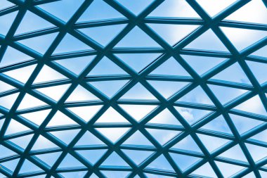 Geometrik metal ve cam çatı yapısının yakın görüntüsü beyaz bulutlu açık mavi gökyüzüne karşı kurulmuş. Görüntü modern mimari ve mühendislik estetiğini vurguluyor.