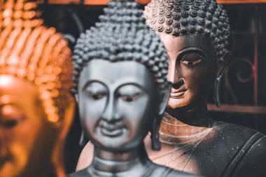 Altın ve gümüş renkli üç Buda heykeli karışık arkaplanlı bir tapınak ortamında bir sırada sergilenmektedir.