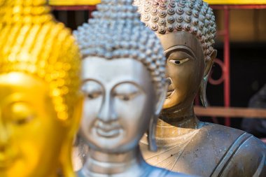 Altın ve gümüş renkli üç Buda heykeli karışık arkaplanlı bir tapınak ortamında bir sırada sergilenmektedir.