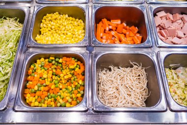 Salata barındaki metal tepsilerde taze salata malzemelerinin karışımı. İçinde mısır, havuç, bezelye, erişte ve doğranmış et var.