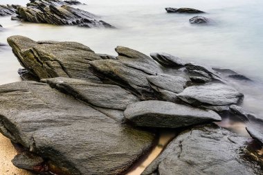 Rocky kıyı şeridinin, bulutlu bir gökyüzünün altında sakin bir denize uzanan pürüzsüz kayalarla uzun süre pozlanması sakin bir kıyı manzarası yaratıyor.