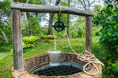 Ahşap bir çerçeve ve makara ile çevrili kırsal bir tuğla su kuyusu bahçedeki yemyeşil yapraklarla çevrili.