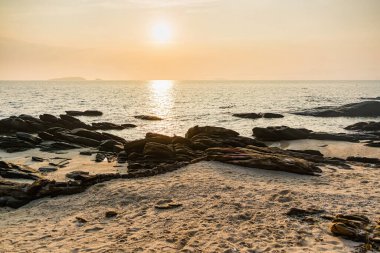 Sarsıntılı kaya oluşumları berrak bir gökyüzünün altında sakin denize doğru uzanır ve sakin bir kıyı manzarası sergiler.