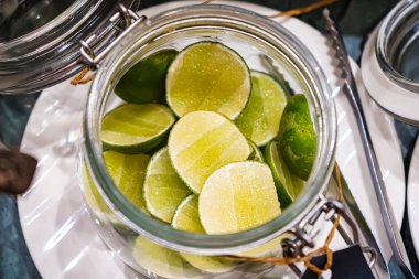 Beyaz bir tabakta dilimlenmiş limonlarla dolu bir kavanoz, yanında metal maşalar ve mavi-yeşil bir arka plan.