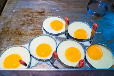 Sıcak ızgaranın üzerinde metal halkalarda pişmiş yumurtalar, parlak sarı sarılar ve yumuşak beyaz bir taban.