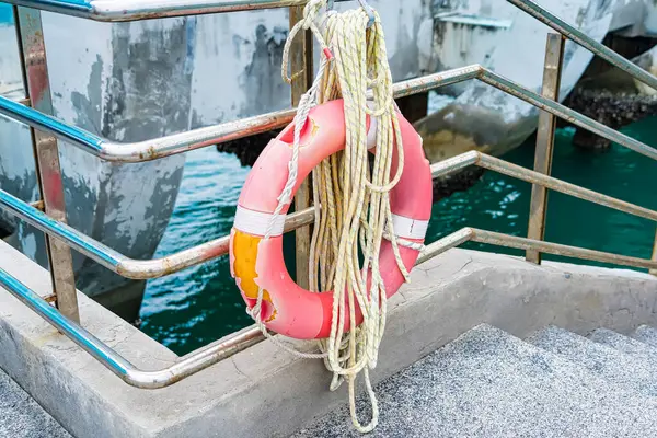 Parlak turuncu yaşam halkası, metal korkuluklarda asılı. Arka planda turkuaz su bulunan rıhtımın kenarında.