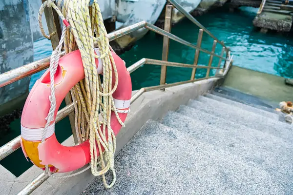 Parlak turuncu yaşam halkası, metal korkuluklarda asılı. Arka planda turkuaz su bulunan rıhtımın kenarında.