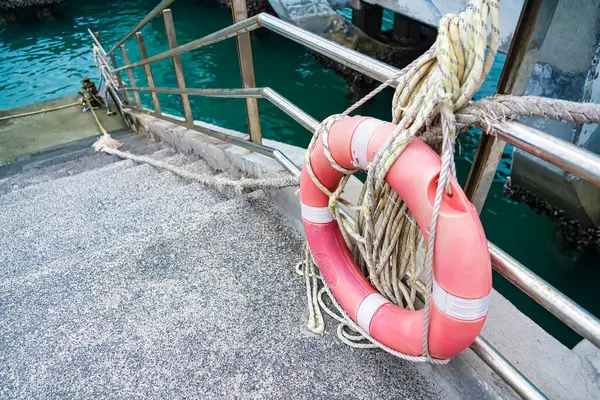 Parlak turuncu yaşam halkası, metal korkuluklarda asılı. Arka planda turkuaz su bulunan rıhtımın kenarında.