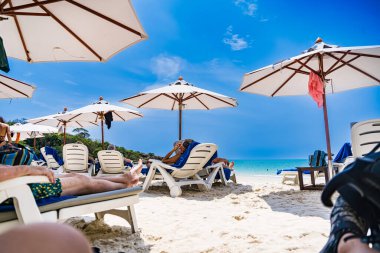 Rayong, Tayland - Nisan 01, 2025: Açık mavi gökyüzü altında sıralanmış renkli güverte sandalyeleri ve şemsiyeleri olan el değmemiş kumlu plaj, Koh Samet, Rayong, Tayland 'da okyanusun tadını çıkaran insanlar