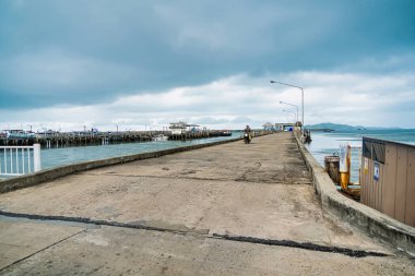 Rayong, Tayland - Nisan 01, 2025 Bulutlu bir gökyüzünün altında uzanan beton iskele, rıhtım boyunca uzanan uzak dağlar ve sokak ışıkları