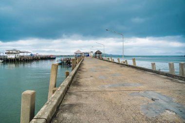 Rayong, Tayland - Nisan 01, 2025 Bulutlu bir gökyüzünün altında uzanan beton iskele, rıhtım boyunca uzanan uzak dağlar ve sokak ışıkları