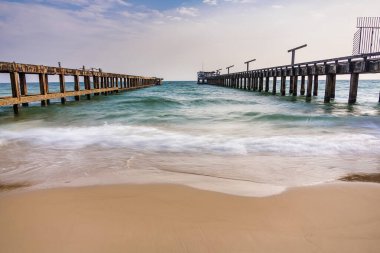 Paralel rıhtımlar okyanusa doğru uzanıyor. Hafif dalgalar kumlu sahile vuruyor. Parçalı bulutlu bir gökyüzünün altında.