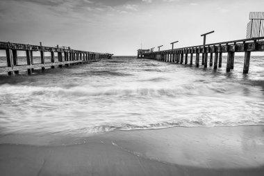 Kırsal paralel rıhtımların uzun süre maruz kaldığı siyah ve beyaz fotoğraf okyanusa doğru uzanıyor. Hafif dalgalar kumlu kumsalda parçalı bulutlu bir gökyüzünün altında süzülüyor.