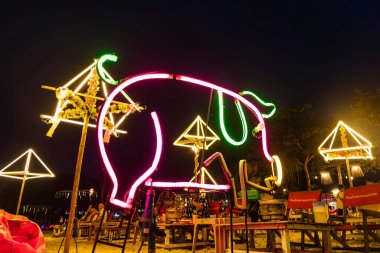 Rayong, Tayland - Nisan 01, 2025: Neon domuz tabelası ve dekoratif aydınlatılmış şemsiyeler, arka planda kırmızı fasulye torbaları üzerinde oturan insanlar