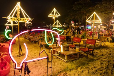 Rayong, Tayland - Nisan 01, 2025: Neon domuz tabelası ve dekoratif aydınlatılmış şemsiyeler, arka planda kırmızı fasulye torbaları üzerinde oturan insanlar
