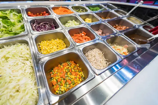 Salata barında, paslanmaz çelik kaplar ve renkli sebzelerle çeşitli taze malzemeler sergileniyor..