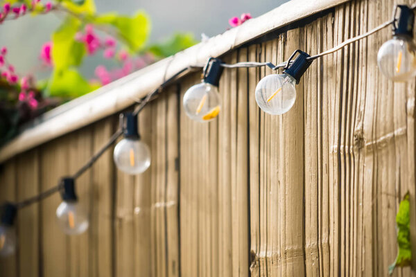 Струнные лампы с обнаженными лампочками накаливания висят на деревянном заборе на открытом воздухе с размытыми цветами на заднем плане