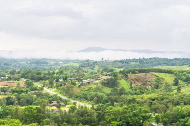 Puslu tepeleri, sık ağaçları ve bulutlu bir gökyüzünün altında dağınık evleri olan yemyeşil vadi manzarası.