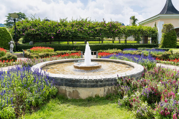 Elegant circular fountain surrounded by colorful flower beds in a beautifully landscaped garden under a partly cloudy sky