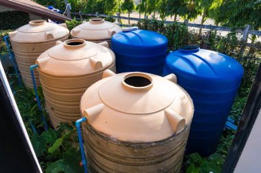 Açık havada yeşil yaprakların arasına yerleştirilmiş ve çevresi bitki örtüsüyle çevrilmiş, kahverengi ve mavi renklerde altı büyük su depolama tankı.