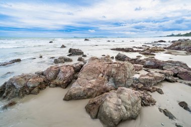 Dağılmış taşlarla kayalık kıyı şeridi ve bulutlu bir gökyüzünün altında alçak gelgit sırasında sakin bir sahil manzarası.
