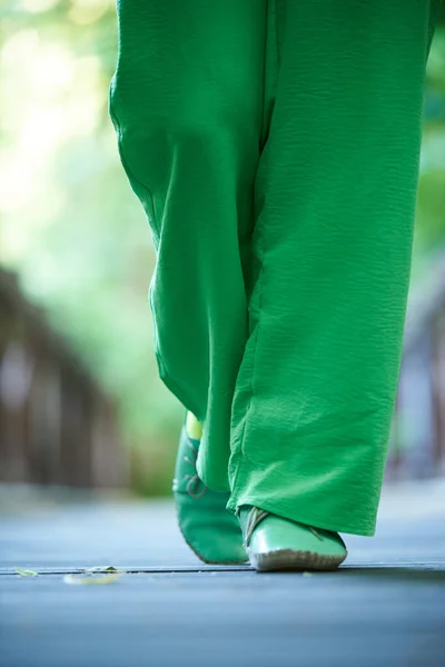 A woman in green trousers and moccasins walks along the road in summer.