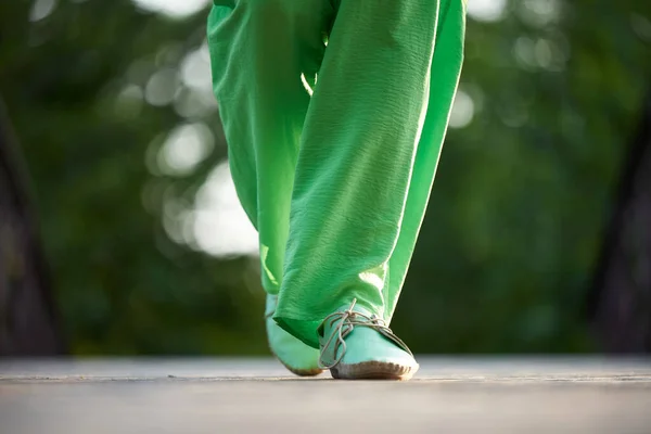 A woman in green trousers and moccasins walks along the road in summer.
