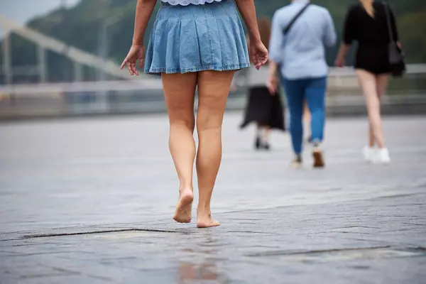 An adult woman in a denim skirt walks barefoot around the city