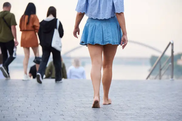 An adult woman in a denim skirt walks barefoot along the city embankment in Kyiv