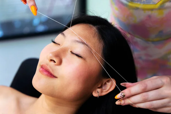 professional beautician performs an eyebrow threading on a young asian woman in the beauty salon, concept of wellness and body care