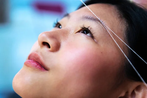 young asian woman having her eyebrows waxed with thread in a beauty salon, wellness and body care concept