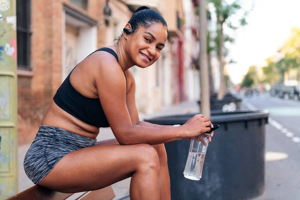 Latina Esportista Segurando Uma Garrafa Água Sorri Enquanto Sentado Olhando — Fotografia de Stock