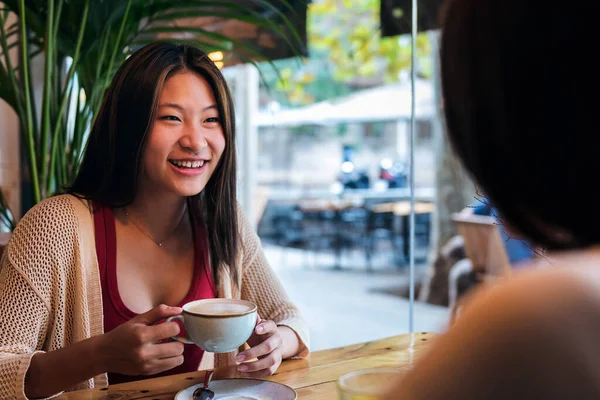 smiling young asian woman drinking a coffee with a friend in a coffee shop, concept of friendship and love between women