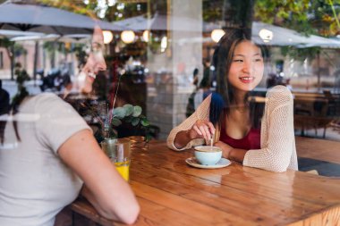 two young friends enjoying having a cup of coffee seen through the glass of the coffee shop, concept of friendship and love between women