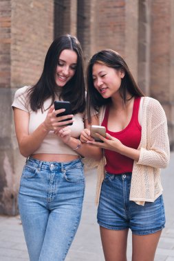 two young women smiling happy looking at their cell phones as they stroll through the city arm in arm during a date, concept of friendship and love between people of the same sex