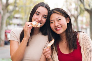 portrait of two smiling women sharing some sweet buns sitting in a city park, concept of friendship and love between people of the same sex