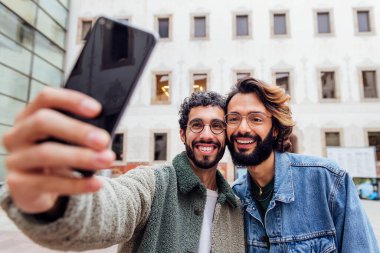 Mutlu bir çift eşcinsel adam sokakta cep telefonuyla selfie çekerken gülüyor. Şehir hayatı ve aynı cinsiyetten insanlar arasında aşk kavramı.