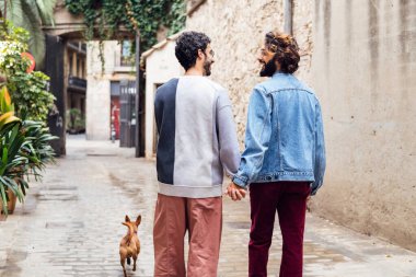 Eşcinsel bir erkek çiftin arka görüntüsü mutlu bir şekilde küçük köpekleri ile birlikte aynı cinsiyetten insanlar arasında bitkilerle, yaşam tarzıyla ve aşkla dolu güzel bir caddede yürüyor.