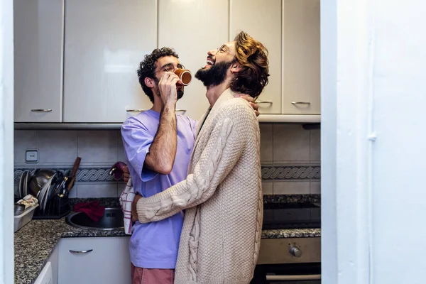 stock image gay male couple hugging and laughing happy while having morning coffee in the kitchen, concept of home lifestyle and love between people of the same sex, copy space for text