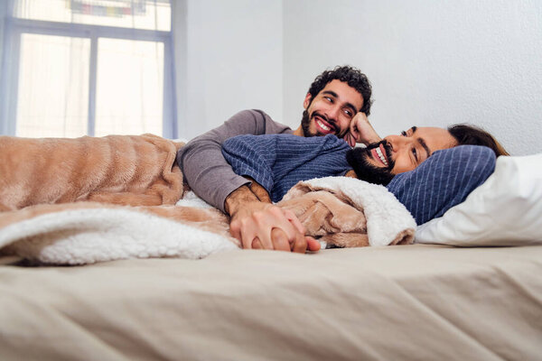 gay male couple relaxing lying on bed with clasped hands, concept of real people lifestyle and love between people of the same sex