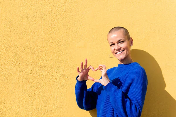 woman with a shaved head and dressed in a blue sweater making a heart shape with her hands against a yellow wall in the background, concept of love and happiness, copy space for text