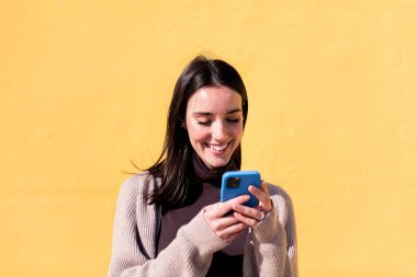 Gülümseyen genç kadın sarı arka planda cep telefonu kullanıyor, iletişim teknolojisi ve modern yaşam tarzı kavramı, metin için kopyalama alanı